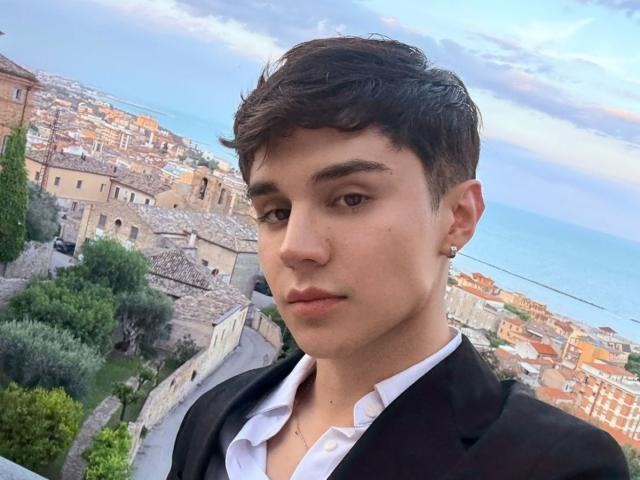 JonyGumm has short brown hair, a nose piercing, and a serious expression. He's wearing a black suit over a white shirt. His ass is not visible.