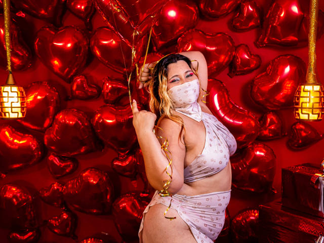 SalmaAzaman with wet blonde hair, wearing a white, polka-dot, halter top and matching skirt, holding gold streamers, standing in front of red heart balloons, masked face, curvy.