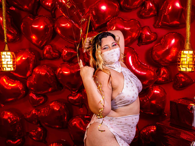 SalmaAzaman with curly blonde hair, white mask, and sheer lingerie, holding gold ribbon, standing against red heart balloons, showing her big ass and small tits, in a sexy pose.