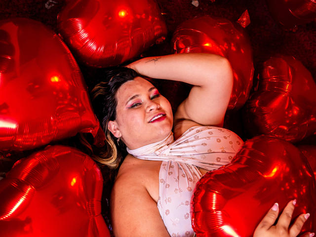 SalmaAzaman with pink eyeshadow, lying among red heart-shaped balloons, smiling, wearing a white halter top, one arm behind head, showing her ass and boobs.