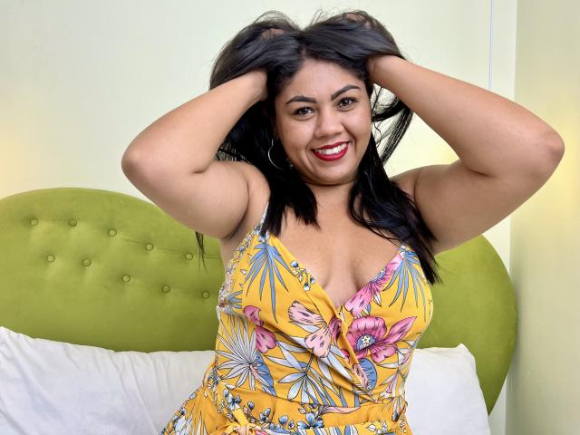 SusanaColls with hands in her black hair, smiling, wearing a yellow floral dress, showing her boobs, red lipstick, hoop earrings, and green headboard in a bright room.