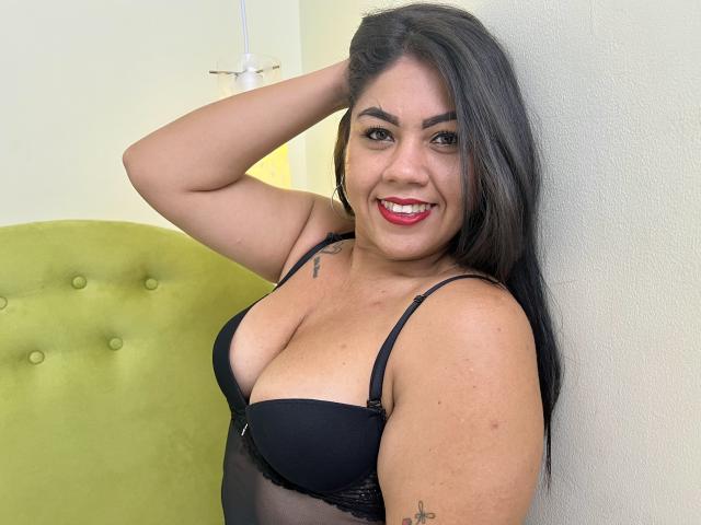 SusanaColls with long black hair, smiling, wearing a black bra, her tits are visible, her hand in her hair, red lipstick, tattoos on her arm, standing against a white wall.