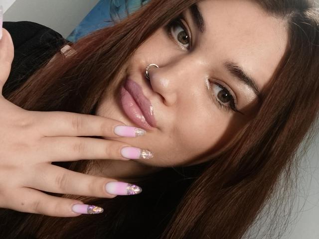 WinterPeggy with brown hair, nose ring, glossy pink lips, and pink nails with gold glitter, touching her face with her hand. Her eyes are brown and she's smiling.