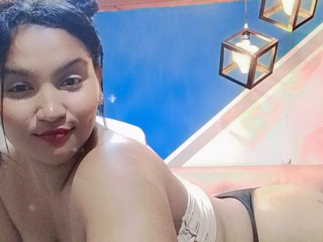 SusanaDiaz, with dark hair in buns, red lipstick, white strapless top, and black thong, laying down, showing her tits and ass, under blue sky with hanging lights.