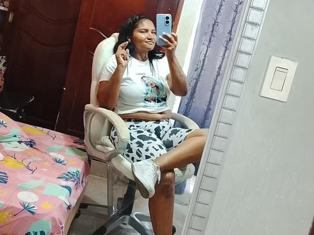 SusanaDiaz with dark skin, medium breasts, wearing a white tee and black/white skirt, sitting in a chair, taking a mirror selfie, middle finger up, in a colorful room.