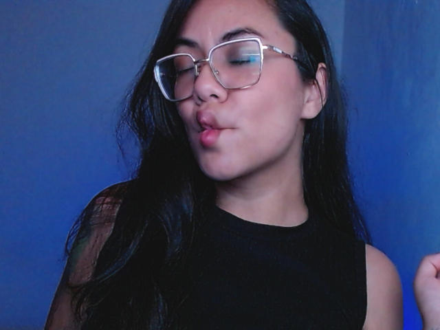 MichellOrtega with glasses, pouting lips, long black hair, black sleeveless top, blue background, looking down, slightly bluish lighting, tattoo on right shoulder.