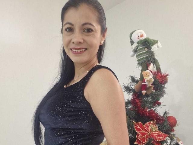 MelissaBaud, with long black hair, wearing a sparkly black sleeveless top, smiling at the camera, standing next to a decorated Christmas tree with a snowman and Santa doll.