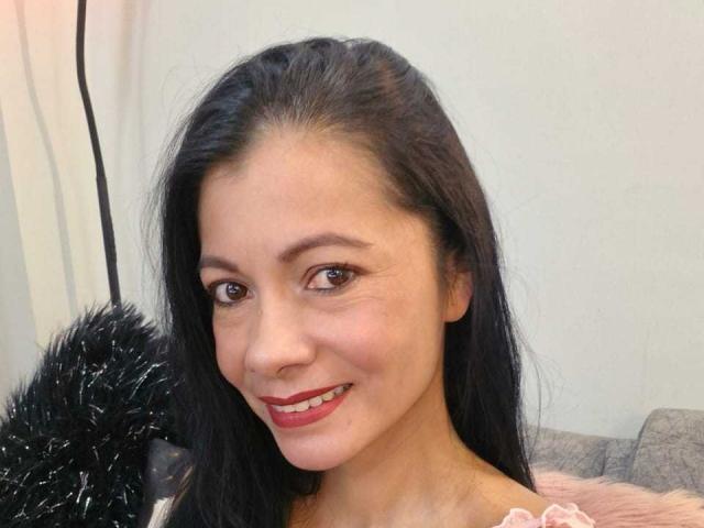 MelissaBaud tiene el pelo largo y negro, ojos marrones y lápiz labial rojo, sonriendo con unos pechos pequeños y lindos, y una blusa rosa, en un selfie. Su nariz está ligeramente levantada..