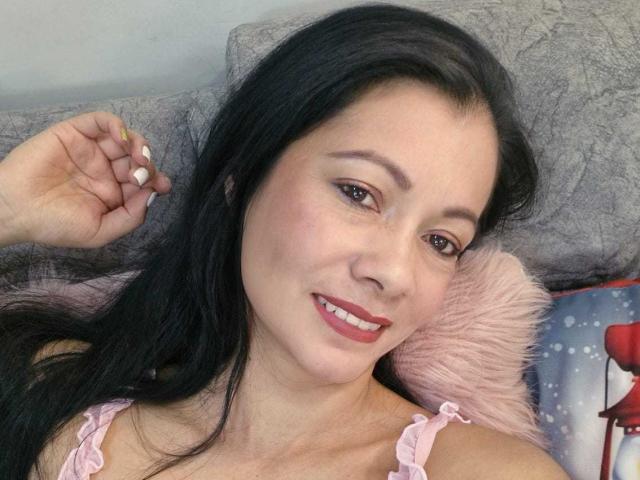 MelissaBaud has black hair, brown eyes, and red lipstick. She's smiling with her hand up, wearing a pink frilled top, showing off her tits. She's lying on a gray pillow.