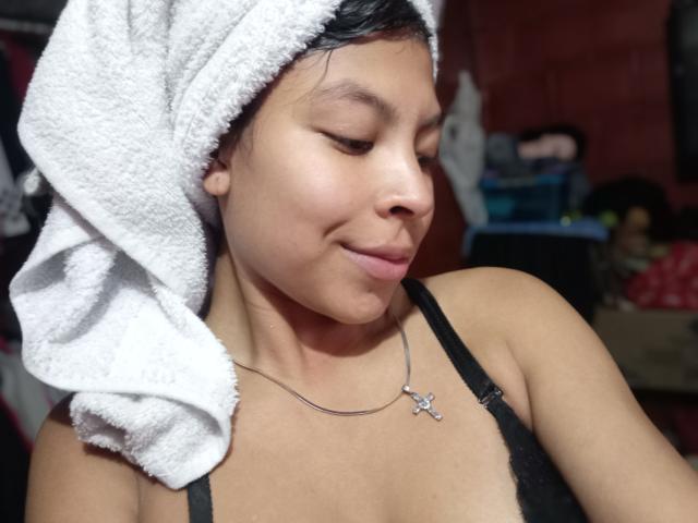 SashaLia has tan skin, black hair wrapped in a white towel, small tits in a black lace bra, and a silver cross necklace. She's looking down with a slight smile.