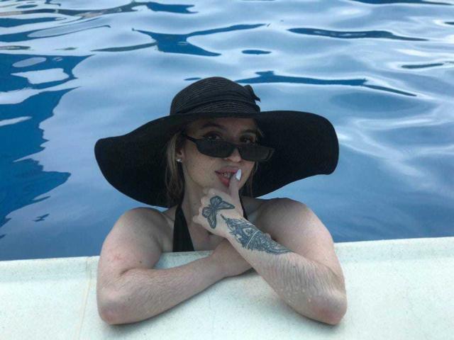 AlinaLee with black sunhat, black sunglasses, and tattoo on right hand, leaning on pool edge, showing her tits above blue water, looking at camera with finger to mouth.