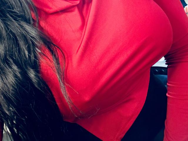 LoveSarahh's red shirt is tight over her big, shiny tits, with long, wavy black hair on the left side. She's wearing black pants, and the focus is on her chest.