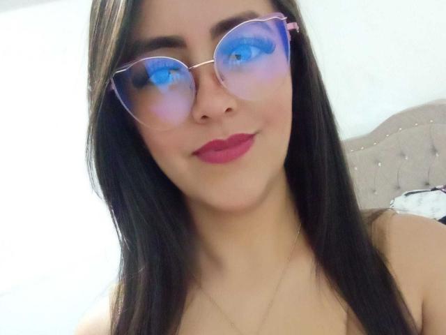 KatiaSmithh with long black hair, wearing pink-tinted round sunglasses and pink lipstick, showing off her boobs slightly in a selfie. She's got a cute smile and a gold necklace.