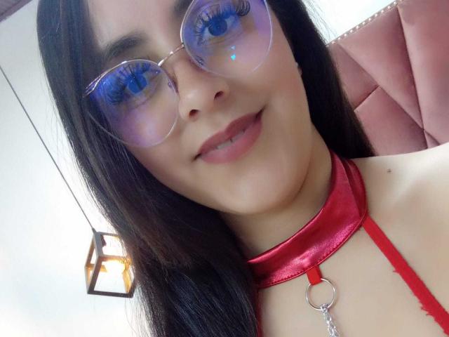KatiaSmithh with long black hair, blue eyes, and round glasses, wearing a red satin choker with a ring and chain, smiling at the camera.