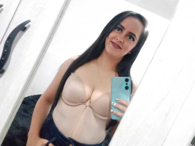 KatiaSmithh with long black hair, beige underboob bra, smiling at mirror, holding blue phone, showing her big tits, in white room, wearing dark pants, looking cute.