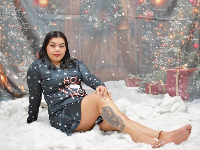 FantasykLadies with black hair, red lipstick, wearing a black snowflake dress, sitting on white fluffy ground, showing tattoo on right thigh, bare feet, Christmas background with trees and gifts.