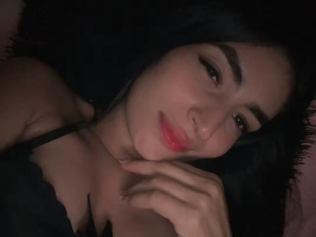 SweetAmandaPolis with black hair, brown eyes, and red lips, lying down, wearing a black lace bra, smiling softly, her hand near her face, in a dimly lit photo.