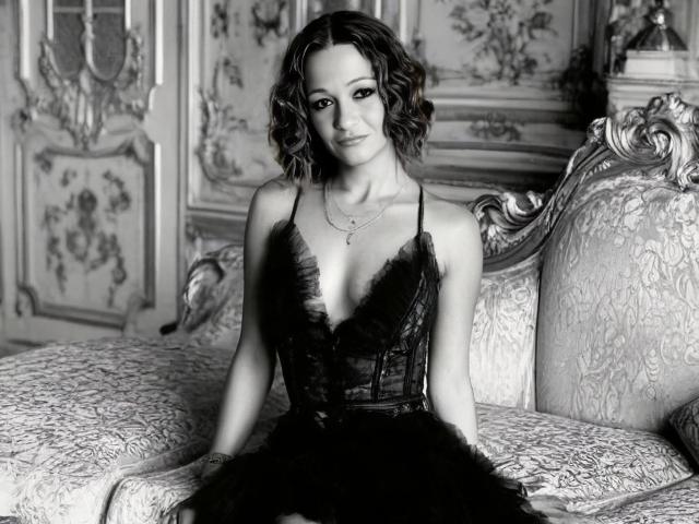 AgathaStone with curly hair, wearing a black lace dress with a deep V, sitting on an ornate bed, showing her tits, in a black-and-white, elegant, vintage-style photo.