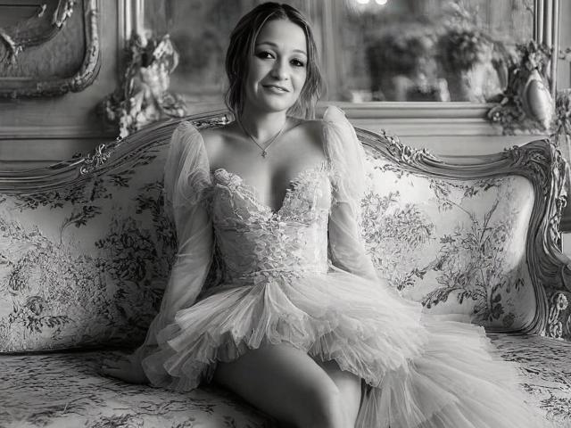 Black-and-white photo of AgathaStone with tits in a lace-up, deep-V, puff-sleeve, tulle dress, sitting on a floral-patterned, ornate couch, smiling softly, legs slightly apart.