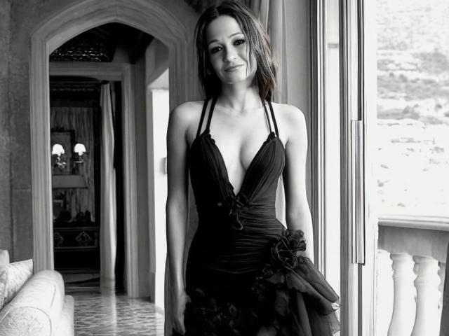 Black-and-white photo of AgathaStone with medium tits in a deep V-neck black dress, standing by a window, smiling, holding a dark floral skirt, and looking cute.