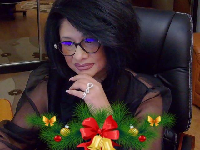 ClassybutNaughty with black hair, glasses, and sheer black top, tongue out, wearing a ring, sitting in a black chair, Christmas wreath with red and yellow bows in front.