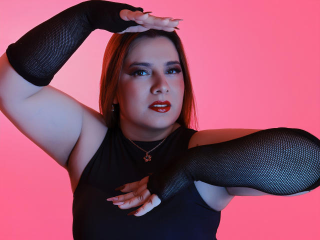 VedaJhonson has red lipstick, black mesh arm sleeves, and a black top, posing with arms raised against a pink background, showing her curvy body and confident expression.