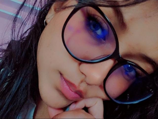 AzulMoon mit ihren großen, blauen Augen, der schwarzen Brille und dem süßen Gesicht, die ihr Kinn in die Hand legt, wobei sie ihre vollen, rosafarbenen Lippen und ihr leicht zerzaustes, dunkles Haar zur Schau stellt..