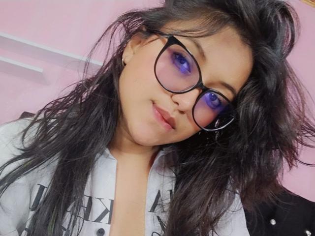 AzulMoon, con el pelo negro despeinado, sombra de ojos morada y gafas, vestida con una camisa blanca, sonriendo ligeramente y mirando a la cámara. Sus pechos son pequeños y su trasero no es visible..