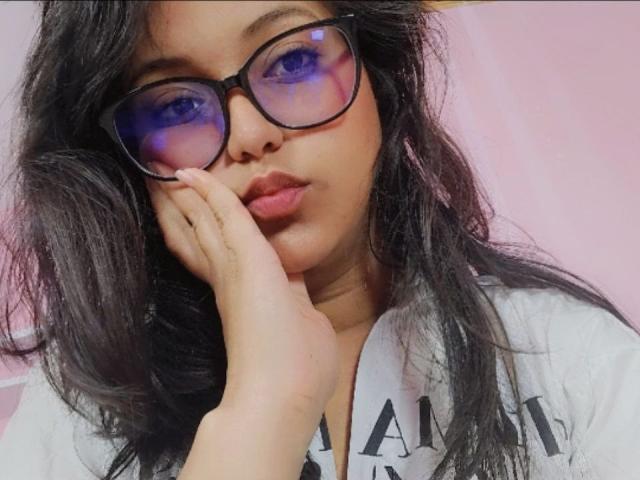 AzulMoon, con gafas de tono azul, cabello negro ondulado y una mano en su mejilla, vestida con una camisa blanca, con una expresión ligeramente aburrida. Sus labios fruncidos son visibles..