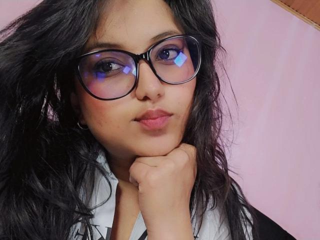 AzulMoon tiene el cabello oscuro y ondulado, usa gafas negras con reflejo azul y apoya su barbilla en la mano. Lleva una camisa blanca con cuello negro..