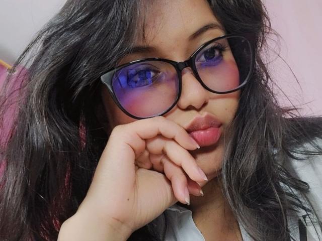 AzulMoon, con el pelo largo y negro, lleva gafas negras con un tono azul, labios rosados y finos, y tiene una mano apoyada en su mejilla. Tiene uñas bonitas, ligeramente desordenadas..