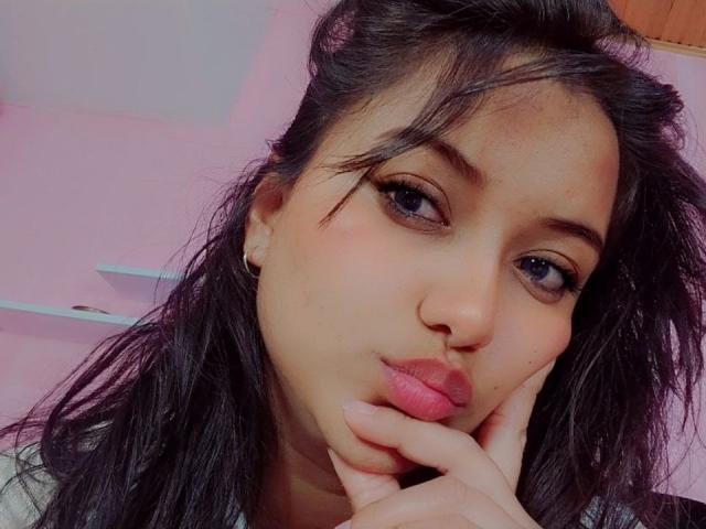 AzulMoon mit pinkem Lippenstift, vollen Lippen, schwarzen Haaren, braunen Augen und einer Hand in der Nähe ihres Mundes. Ihre Nase ist klein, und sie trägt schwarze Ohrstecker..
