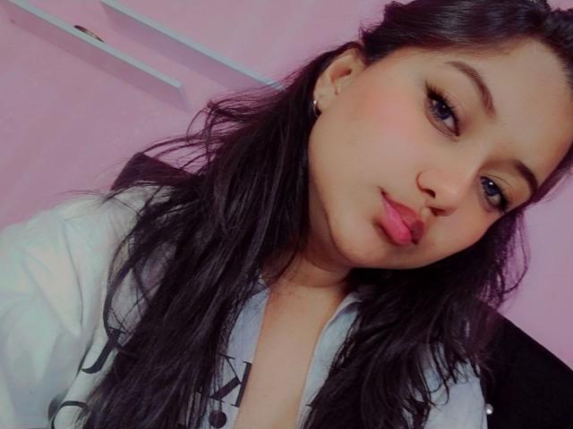 AzulMoon has long black hair, light brown skin, and is pouting with pink lips. She wears a white shirt, has small earrings, and looks directly at the camera.