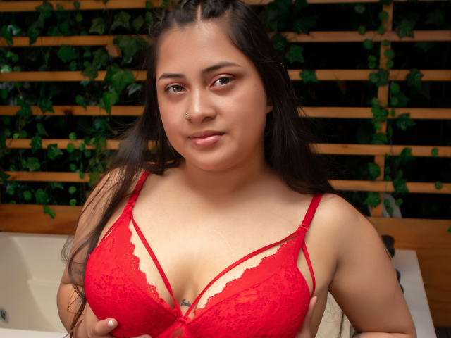 HannaSkyler with medium skin, long black hair, and nose ring, wearing a red lace bra, showcasing her large, round boobs, and a small tattoo below her cleavage.