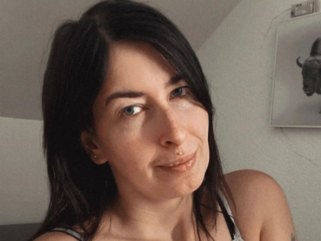 Candismimi-hot has dark brown hair, green eyes, a nose piercing, and a slight smile. She's wearing a gray top, showing her bare shoulders. She's cute and sexy.