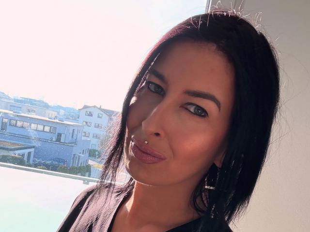Candismimi-hot has long black hair, green eyes, and a nose piercing. She's pouting with pink lips, wearing a black top. Sunlight highlights her tan skin.