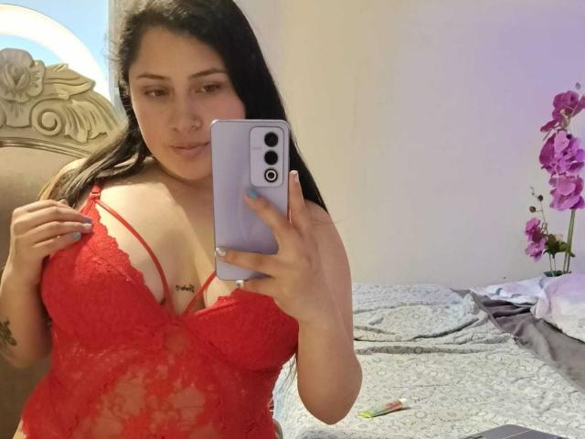 HannaSkyler with long black hair, wearing red lace lingerie, showing her tits in a mirror selfie, holding a purple phone, with a pink orchid in the background.