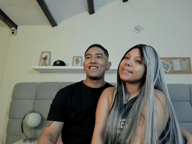 EroticXSins with short black hair, black t-shirt, and tattoo on arm, sits beside EroticXSins with long silver hair, sleeveless top, smiling at camera. Both have cute, happy expressions.