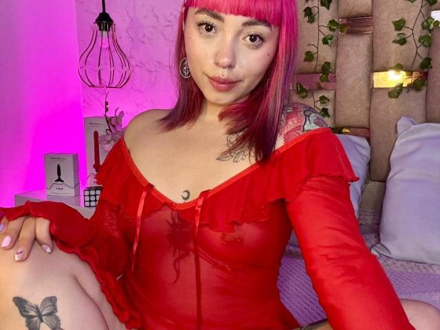 GigiVolkova with pink hair, nose ring, and tattoos, wearing a sheer red off-shoulder top, showing small tits and a butterfly tattoo on her thigh.