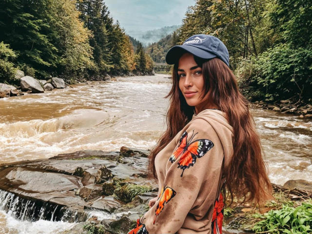 MandiBrooks with long brown hair, blue cap, butterfly-patterned hoodie, standing by a rocky, flowing river, looking over her shoulder, smiling, green forest in background.