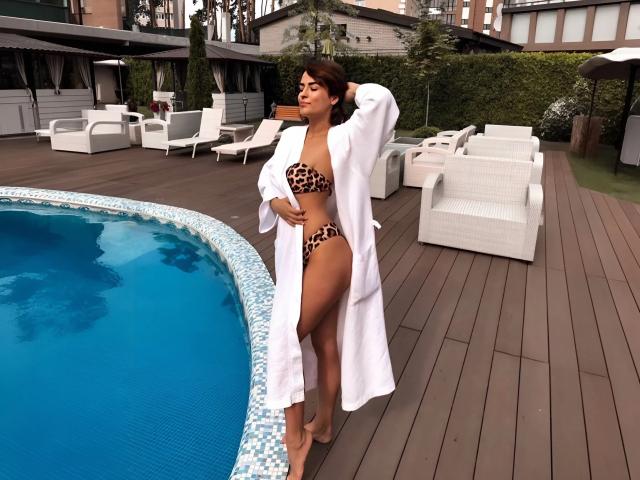 MandiBrooks with tan skin, brown hair, and a curvy figure, wearing a leopard print bikini and white robe, standing by a blue-tiled pool, hand on hip, looking sexy.
