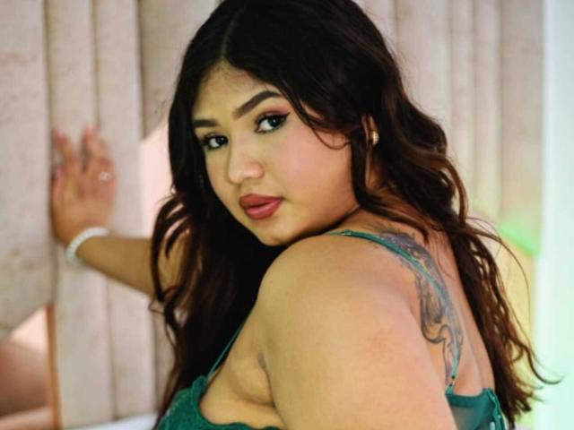 SofyaZaens with wavy dark hair, green dress, looking over shoulder, tattoo on right shoulder, hand on wooden wall, green and white light, red lipstick, black eyeliner.