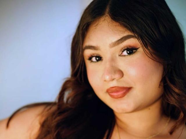 SofyaZaens has dark brown wavy hair, full pink lips, and cute eyeliner. Her skin is tan, and she's smiling softly at the camera. Her tits are partially visible.