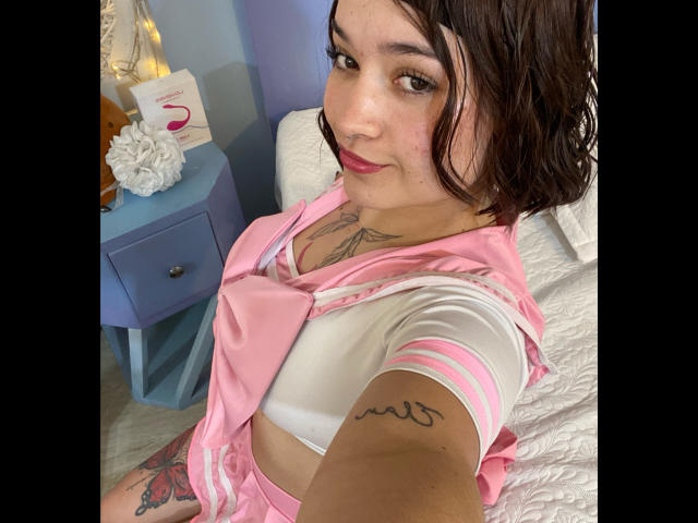 AnikaFleur with short brown hair, pink and white schoolgirl outfit, tattoos on chest and arm, red lipstick, white bed, blue nightstand with white bag and flowers.