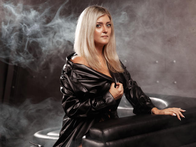 HelenUnique with blonde hair, wearing a shiny black leather jacket, exposing her tits, leaning on a black leather couch, surrounded by wispy smoke. She looks confident.