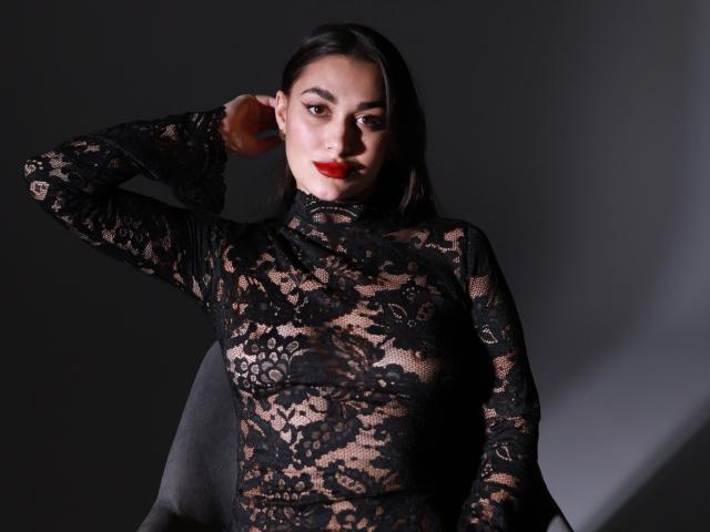 BrunetteBabeXX-hot with dark hair, red lipstick, and black lace dress, showing tits through lace, hand in hair, sitting, grey background, looking at camera.