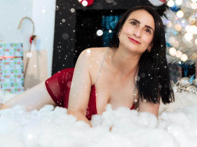 BrendaCantorel, with black hair and red dress, lying in white snow, showing her tits, smiling, silver chain necklace, Christmas tree lights in the background, snowflakes falling.