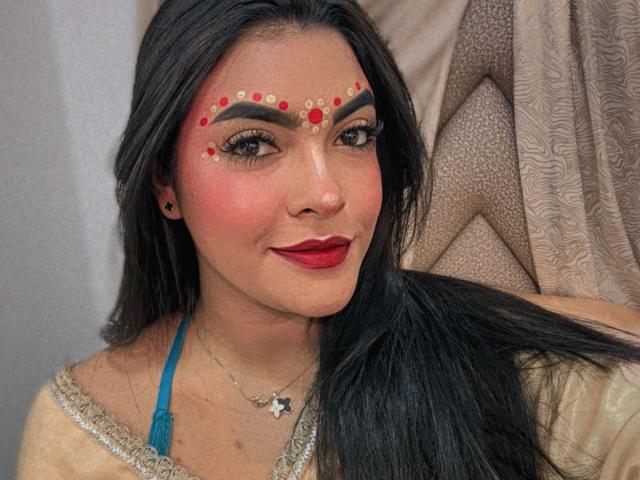 LakshmIrani has long black hair, red lipstick, and intricate facial makeup with red dots and gold patterns. She's wearing a blue halter top and a silver necklace.