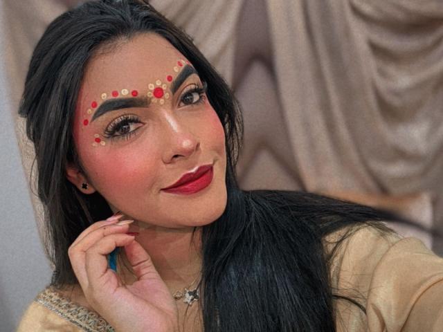 LakshmIrani has dark hair, red lipstick, and bold makeup with red and gold patterns on her forehead. She's smiling with her hand near her face.