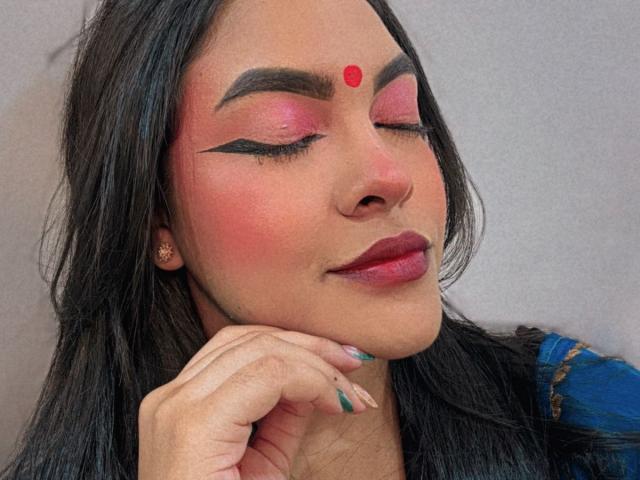 LakshmIrani has pink eyeshadow, black winged eyeliner, red dot on forehead, pink blush, red lipstick, black hair, blue top, gold earrings, and teal nail polish, smiling with eyes closed.